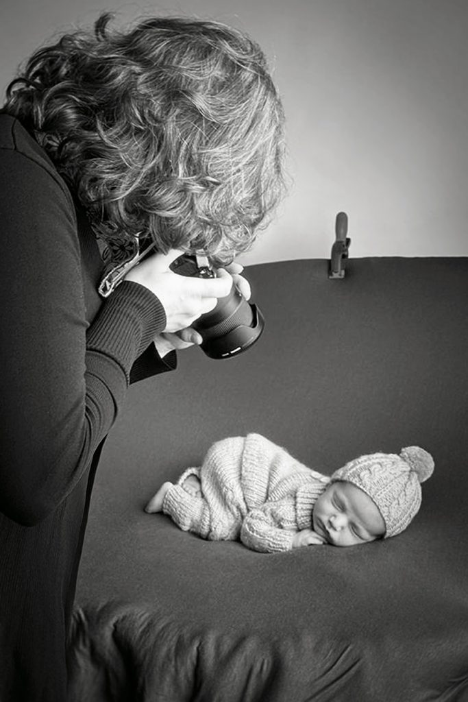 Un fotografo tiene in mano una macchina fotografica e cattura momenti preziosi di un neonato addormentato. La scena, catturata da Tania Ayyad Photography durante il servizio newborn a Palermo.
